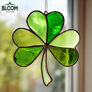 May include: A stained glass shamrock suncatcher in shades of green. The cloverleaf design is outlined in a dark brown metal. The Bloom Vector logo is in the upper left corner. The suncatcher is suspended in front of a window.