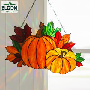 May include: Stained glass pumpkin and autumn leaf decoration. The pumpkins are orange and yellow, with brown stems. The leaves are red, orange, and green. The decoration hangs from a chain and is illuminated by sunlight.