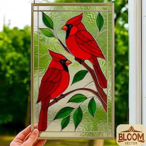 May include: Stained glass panel featuring two vibrant red cardinal birds perched on a brown branch with green leaves. The birds have black masks and orange beaks. The panel is framed in a gold-colored metal and has the text "BLOOM VECTOR".