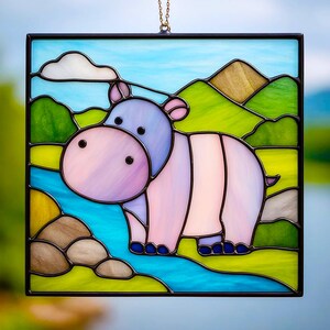 Peut inclure: Panneau en vitrail représentant un hippopotame fantaisiste dans des tons roses et violets, sur un paysage de collines vertes, une rivière bleue et un ciel bleu clair avec un nuage blanc. Le panneau est encadré en noir et suspendu par une chaîne dorée.