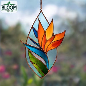 Op de afbeelding: Gebrandschilderde zonnevanger in de vorm van een paradijsvogelbloem. Het ontwerp bevat oranje, blauwe en groene glazen stukken, omlijnd met koperkleurig metaal. De zonnevanger hangt aan een fijne ketting, met het "Bloom Vector" logo linksboven.