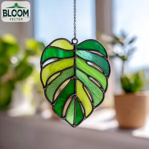 Op de afbeelding: Gebrandschilderd glazen suncatcher in de vorm van een monstera blad, in groen- en geeltinten. Het blad is omlijst door een donker metalen frame en hangt aan een ketting. Het Bloom Vector logo is linksboven zichtbaar.