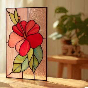 May include: Stained glass panel featuring a vibrant red hibiscus flower with pink and coral petals, green and yellow leaves, and a black frame. The floral design is set against a textured, light pink background, creating a decorative art piece.