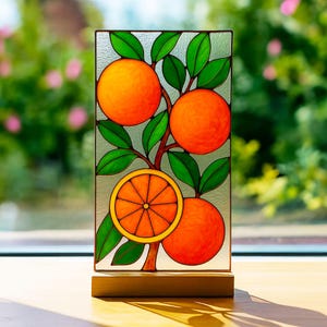 Op de afbeelding: Een glas-in-loodpaneel met een sinaasappelboom met drie hele sinaasappels en een doorgesneden sinaasappel. De sinaasappels zijn fel oranje, de bladeren zijn groen en de takken zijn bruin. Het paneel staat op een houten voet.