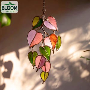 Pode incluir: Uma decoração suspensa em vitral com folhas em forma de coração em tons de rosa, laranja e verde. As folhas são conectadas por uma moldura de metal e suspensas por uma corrente. O logotipo Bloom Vector está no canto superior esquerdo.