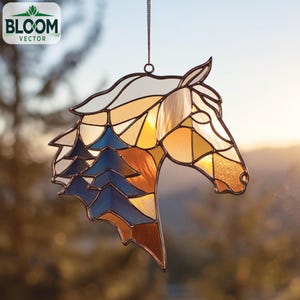 Puede incluir: Atrapasoles de cabeza de caballo de vidriera con una cadena para colgar. El diseño presenta una cabeza de caballo con una melena y un paisaje de árboles azules. El vidrio es una mezcla de blanco, amarillo, naranja y marrón.