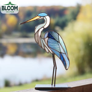 May include: A stained glass heron sculpture with a blue, white, and gray color scheme. The bird has a long yellow beak and stands on a metal base. The Bloom Vector logo is in the upper left corner.