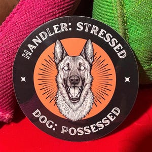 May include: Round sticker with a black border and the text "HANDLER: STRESSED DOG: POSSESSED". The sticker features a detailed illustration of a German Shepherd dog with glowing white eyes and an open mouth, set against an orange burst.