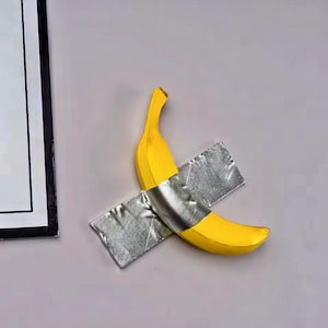 Könnte beinhalten: Eine reife gelbe Banane ist mit zwei Stücken silbernem Klebeband an einer hellgrauen Wand befestigt. Die Banane ist diagonal positioniert und bildet einen auffälligen Kontrast zum neutralen Hintergrund.