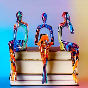 May include: Three abstract sculptures of seated figures with colourful, swirling paint patterns. The figures are perched on a stack of books. The sculptures feature a variety of colours, including blue, orange, red, and yellow.
