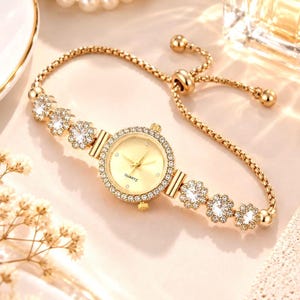 May include: A gold-tone quartz wristwatch featuring a round face with a gold dial and a bezel encrusted with clear crystals. The bracelet consists of a delicate gold chain adorned with six flower-shaped crystal charms and an adjustable sliding clasp.