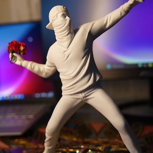 Peut inclure: Figurine blanche représentant une personne en sweat à capuche, casquette et cagoule, tenant un bouquet de fleurs rouges. La figurine est dans une pose dynamique, un bras tendu. L'arrière-plan est flou.