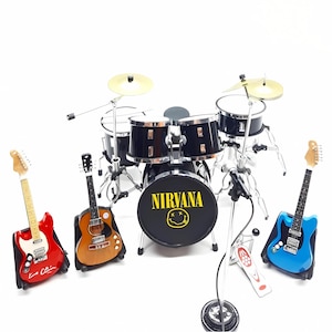 Może przedstawiać: Miniaturowy czarny zestaw perkusyjny ze złotymi talerzami, mikrofonem i trzema gitarami w kolorze czerwonym, brązowym i niebieskim. Na bębnie basowym napis "NIRVANA" i uśmiechnięta buzia.