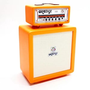 Könnte beinhalten: Ein orangefarbenes Gitarrenverstärker- und Lautsprecherbox-Set. Der Verstärker mit der Aufschrift "Orange" steht auf der Lautsprecherbox. Die Lautsprecherbox hat eine weiße Lautsprecherabdeckung mit dem "Orange"-Logo. Der Verstärker hat Knöpfe und einen Griff.