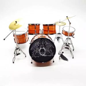 May include: Miniature drum set with a Queen logo on the bass drum. The set includes a bass drum, snare, two toms, a hi-hat, and a cymbal. The drums have a natural wood finish and are mounted on silver stands.