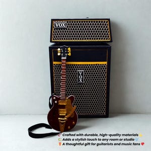 Op de afbeelding: Een miniatuur gitaar- en versterkerset. De gitaar is bruin en goudkleurig, met een zwarte band. De versterker is zwart met gouden randen en een luidsprekerrooster. Het woord "VOX" is zichtbaar.