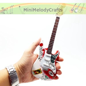 May include: Miniature electric guitar with a white body, red accents, and decorative floral and scroll designs. The guitar has a brown neck and a silver bridge and strings. The headstock is light-colored. The guitar is held in a hand.