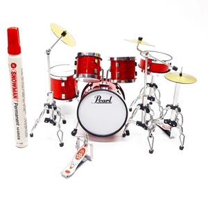 May include: A miniature red drum kit with silver stands and yellow cymbals. The kit includes a bass drum, snare drums, and a hi-hat. A red and white Snowman permanent marker is next to the drum kit.