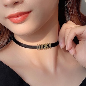 Może przedstawiać: Czarny naszyjnik typu choker z napisem "LUCKY" w złotych literach. Naszyjnik wykonany jest z materiału przypominającego aksamit i przylega blisko szyi. Zdjęcie przedstawia zbliżenie naszyjnika.