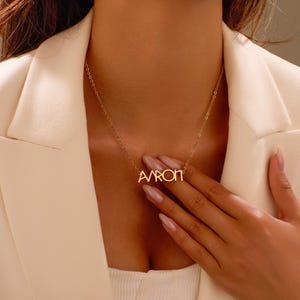 Puede incluir: Un collar dorado con el nombre "AARON" en una fuente cursiva delicada. El collar se muestra sobre una blazer color crema. La cadena es fina y el colgante est&aacute; centrado.