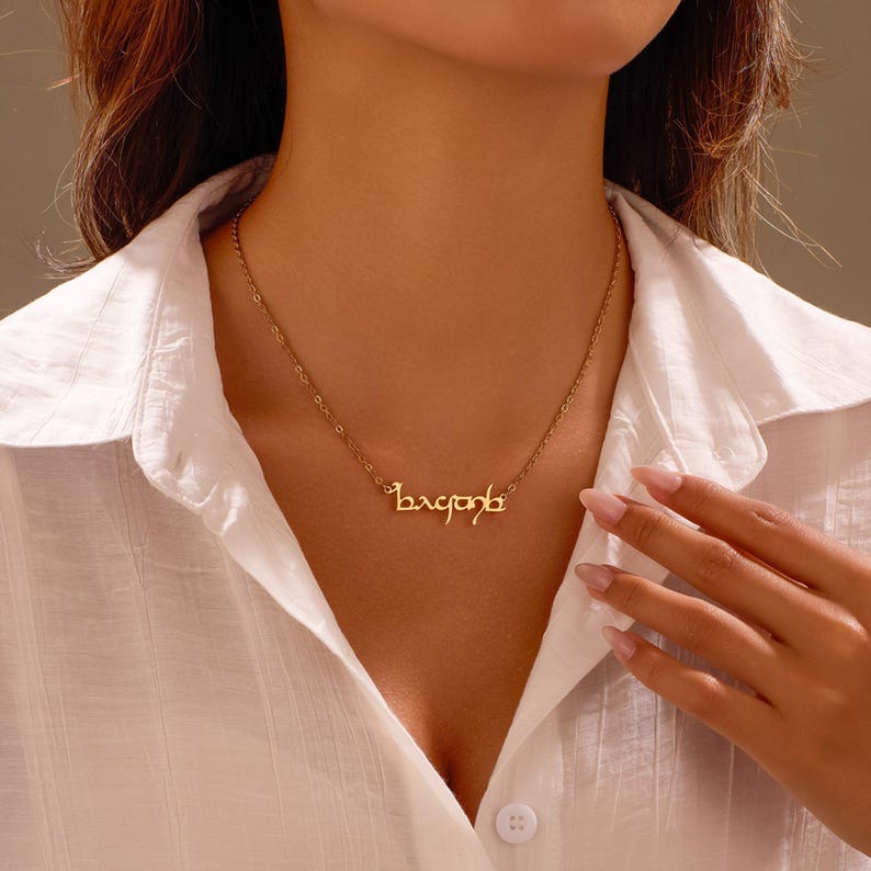 Puede incluir: Un collar dorado con una cadena delicada y un colgante con texto estilizado. El collar se lleva sobre una camisa blanca con cuello. El texto del colgante est&aacute; en escritura cursiva.