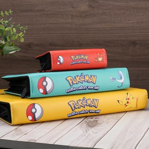May include: Three colorful Pokemon card binders stacked on top of each other. The top binder is red, the middle one is teal, and the bottom one is yellow. Each binder has the Pokemon logo and the phrase "Gotta catch 'em all!"