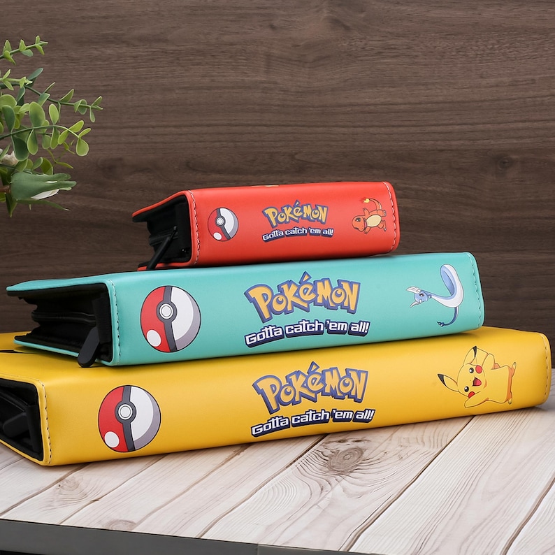 May include: Three colorful Pokemon card binders stacked on top of each other. The top binder is red, the middle one is teal, and the bottom one is yellow. Each binder has the Pokemon logo and the phrase "Gotta catch 'em all!"