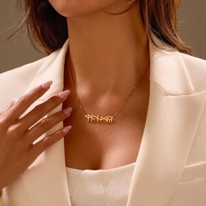 Puede incluir: Un collar dorado con un colgante con letras estilizadas. El collar se lleva sobre una blazer blanca. El dise&ntilde;o del colgante es el punto central, mostrando una est&eacute;tica &uacute;nica y elegante. La cadena es delicada, complementando el dise&ntilde;o del colgante.