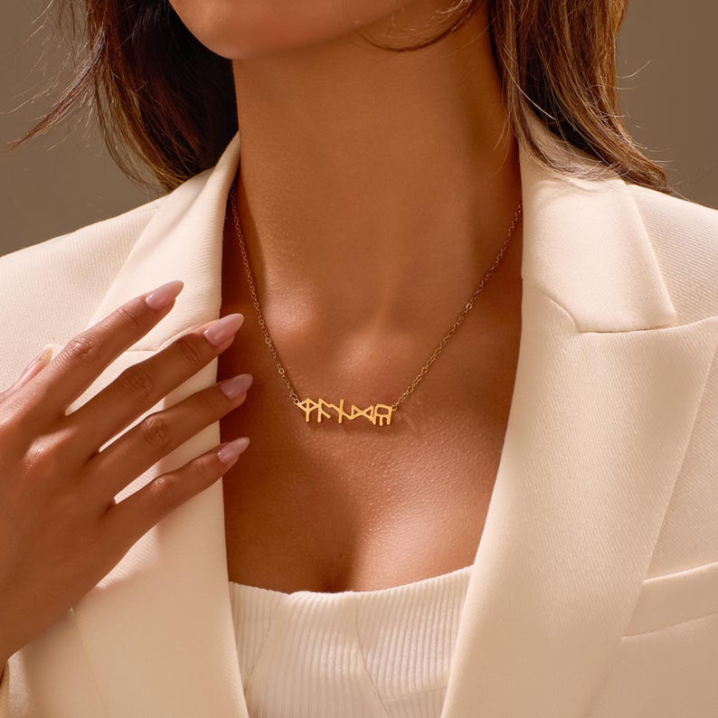 Puede incluir: Un collar dorado con un colgante con letras estilizadas. El collar se lleva sobre una blazer blanca. El dise&ntilde;o del colgante es el punto central, mostrando una est&eacute;tica &uacute;nica y elegante. La cadena es delicada, complementando el dise&ntilde;o del colgante.