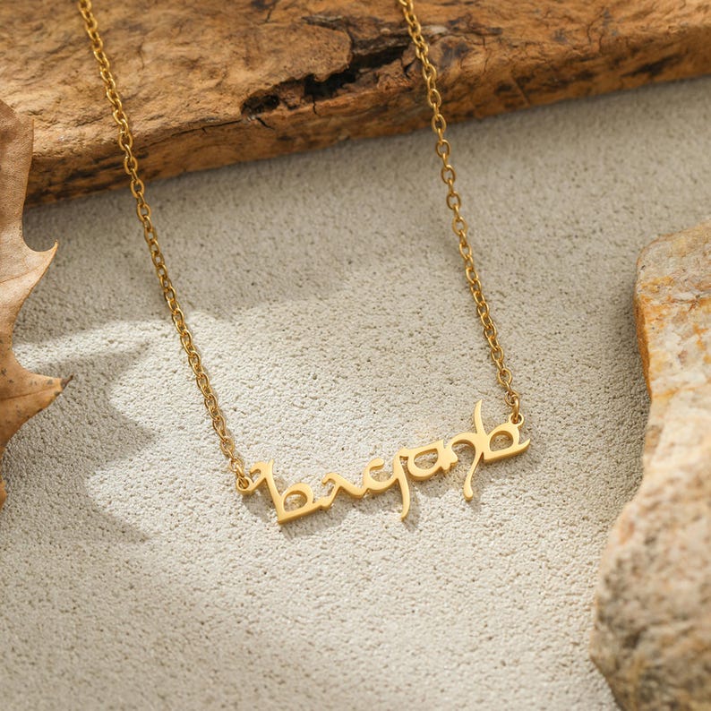 Puede incluir: Un collar dorado con el nombre "bryanb" en una escritura estilizada. El collar se muestra sobre una superficie clara, con un fondo de madera y algunas piedras.