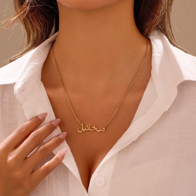 Puede incluir: Un collar dorado con una cadena delicada. El colgante presenta escritura &aacute;rabe. El collar se lleva sobre una camisa blanca abotonada.