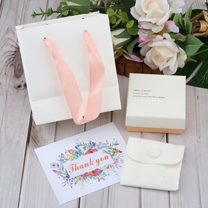 Puede incluir: Una bolsa de regalo blanca con asas de cinta rosa, una peque&ntilde;a caja de joyer&iacute;a, una tarjeta con el texto "Thank you" y una peque&ntilde;a bolsa. La tarjeta tiene un dise&ntilde;o floral. La caja de joyer&iacute;a tiene el texto "SMALL JEWELRY unique beauty only belong to the best of you."