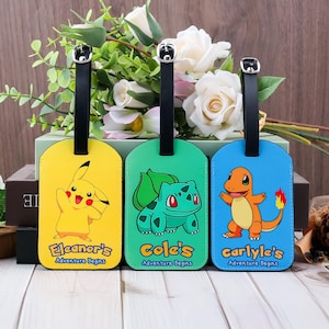 May include: Three colourful luggage tags featuring cartoon characters. The yellow tag has Pikachu, the green tag has Bulbasaur, and the blue tag has Charmander. Each tag has a black strap and the text "Adventure Begins".