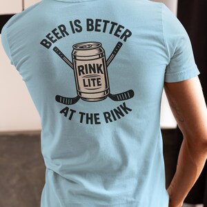 Puede incluir: Camiseta azul claro con la frase "BEER IS BETTER AT THE RINK" en la espalda. El diseño incluye una lata de cerveza y palos de hockey cruzados. La camiseta tiene cuello redondo y mangas cortas.