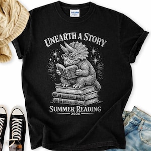 May include: A black short-sleeve t-shirt featuring a white graphic of a triceratops wearing glasses and reading a book while sitting on a stack of books. The text reads UNEARTH A STORY and SUMMER READING 2026 in a distressed white font.