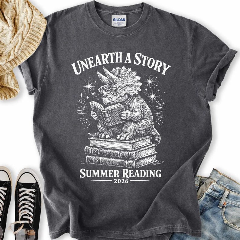 May include: A charcoal gray short-sleeve t-shirt featuring a white graphic of a triceratops wearing glasses and reading a book while sitting on a stack of books. The text reads UNEARTH A STORY SUMMER READING 2026 in a white serif font.