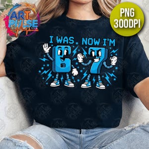 May include: Black t-shirt with a blue graphic that reads "I WAS. NOW I'M" above cartoon numbers 6 and 7. The numbers are blue with white accents and lightning bolt details. The shirt has a round neckline.