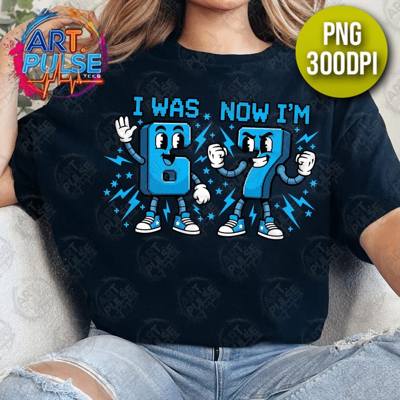 May include: Black t-shirt with a blue graphic that reads "I WAS. NOW I'M" above cartoon numbers 6 and 7. The numbers are blue with white accents and lightning bolt details. The shirt has a round neckline.