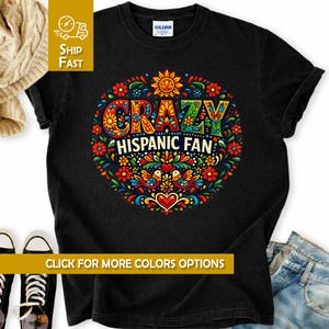 Può includere: T-shirt nero con un design floreale colorato e la scritta "CRAZY HISPANIC FAN". Il design include un sole, uccelli e un cuore. La maglietta è realizzata in un materiale morbido.