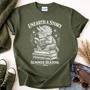 May include: An olive green t-shirt featuring a white graphic of a triceratops wearing glasses and reading a book while sitting on a stack of books. The text reads "UNEARTH A STORY" above the dinosaur and "SUMMER READING 2026" below the stack.