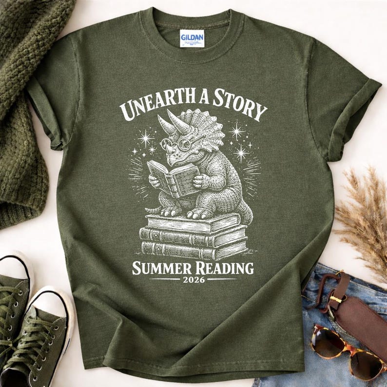 May include: An olive green t-shirt featuring a white graphic of a triceratops wearing glasses and reading a book while sitting on a stack of books. The text reads "UNEARTH A STORY" above the dinosaur and "SUMMER READING 2026" below the stack.
