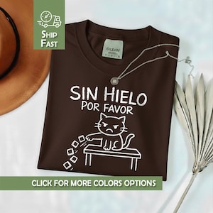 Könnte beinhalten: Ein braunes T-Shirt mit dem Text "SIN HIELO POR FAVOR" und einer Cartoon-Katzen-Illustration. Die Katze sitzt auf einem Tisch und scheint Eiswürfel wegzuschieben. Eine silberne Halskette liegt über dem Shirt.