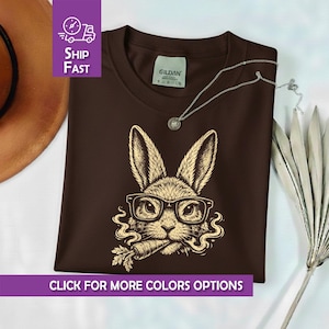 Könnte beinhalten: Ein braunes T-Shirt mit einer detaillierten Illustration eines Hasen mit Brille und einer Karotte. Das Hasen-Design ist in einem hellbraunen Farbton gehalten. Das Shirt trägt den Text "CLICK FOR MORE COLORS OPTIONS".