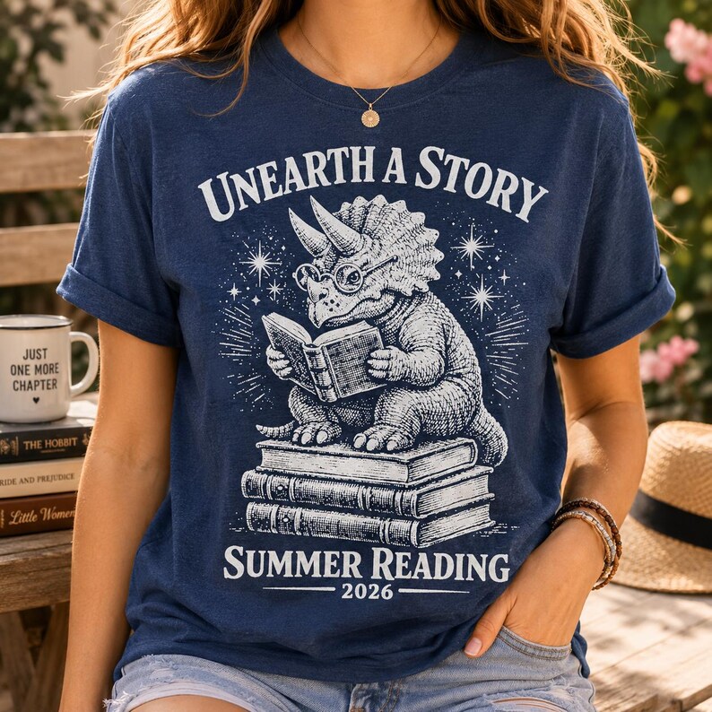 May include: A navy blue short-sleeve t-shirt featuring a white graphic of a triceratops wearing glasses and reading a book while sitting on a stack of three books. The text reads "UNEARTH A STORY" at the top and "SUMMER READING 2026" at the bottom.