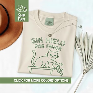 Könnte beinhalten: Cremefarbenes T-Shirt mit grünem Text und Katzenillustration. Der Text lautet "SIN HIELO POR FAVOR". Die Illustration zeigt eine Katze, die einen Becher mit Eiswürfeln umwirft. Eine silberne Halskette liegt auf dem Shirt.