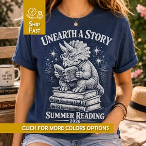 May include: A navy blue t-shirt featuring a white graphic of a triceratops wearing glasses and reading a book while sitting on a stack of books. The text reads UNEARTH A STORY SUMMER READING 2026. The design includes small star-like accents.