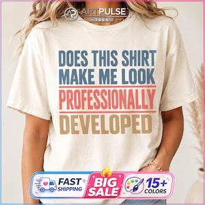 May include: A cream-colored short-sleeve t-shirt featuring the text "DOES THIS SHIRT MAKE ME LOOK PROFESSIONALLY DEVELOPED" printed in blue, red, and tan block letters. The shirt has a crew neckline and a relaxed fit with a standard hemline.