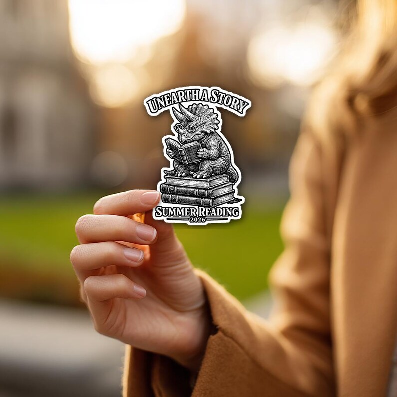 May include: A die-cut sticker featuring a black and white illustration of a triceratops wearing glasses and reading a book while sitting on a stack of books. The text reads UNEARTH A STORY at the top and SUMMER READING 2026 at the bottom.