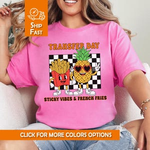 Puede incluir: Camiseta rosa con un gráfico de una piña de dibujos animados con gafas de sol en forma de corazón y una caja de patatas fritas. El texto "TRANSFER DAY" y "STICKY VIBES & FRENCH FRIES" también está impreso.