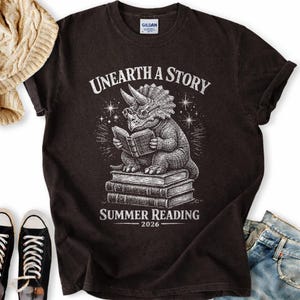 May include: A black short-sleeve t-shirt featuring a white graphic of a triceratops wearing glasses and reading a book while sitting on a stack of books. The text reads "UNEARTH A STORY" above and "SUMMER READING 2026" below the illustration.